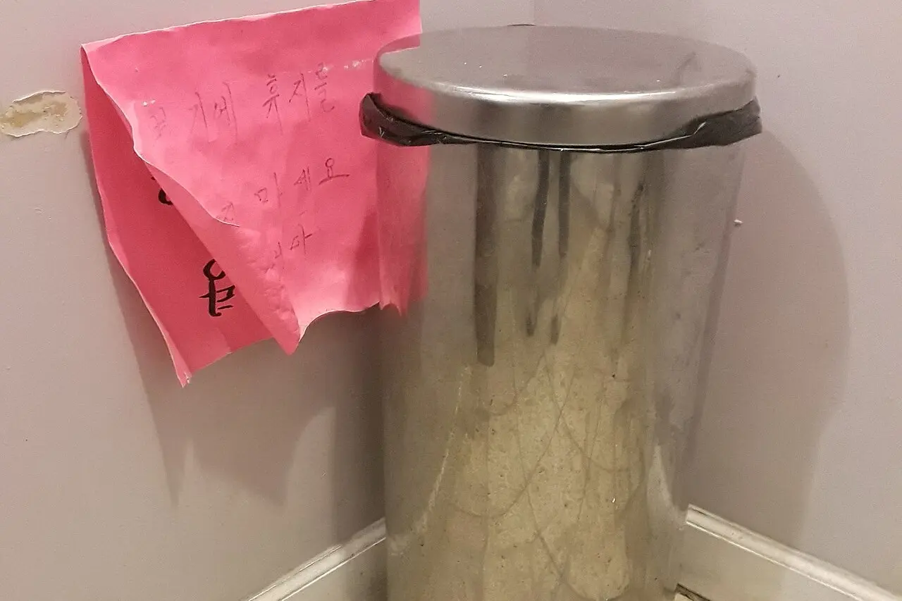 Lixeira de inox com a tampa fechada, no canto de um banheiro, ao lado de um aviso de papel rosa colado na parede.
