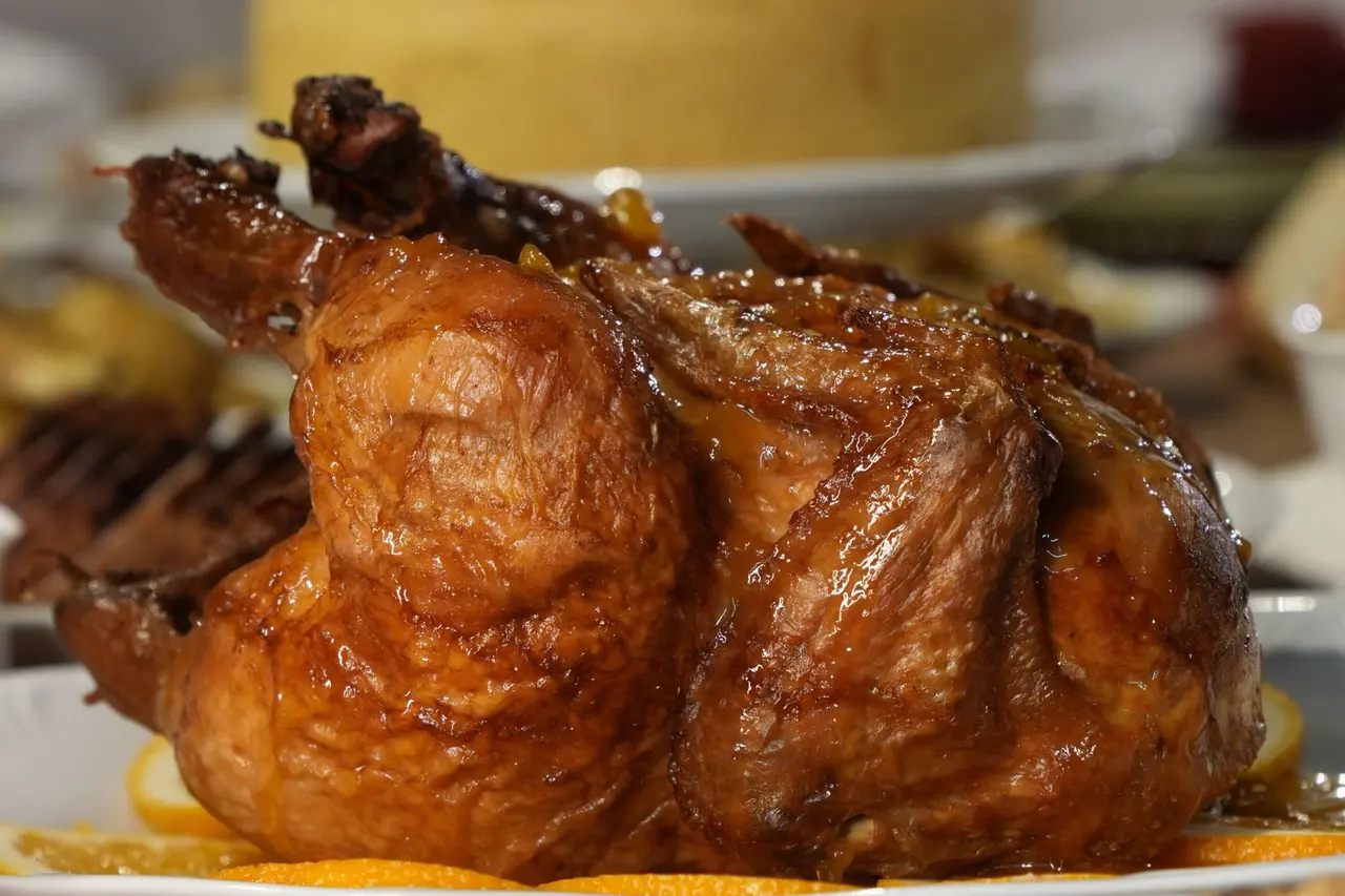 Ave assada inteira, de pele bem dourada, numa travessa com rodelas de laranja, com um bolo e outros pratos de festa ao fundo.