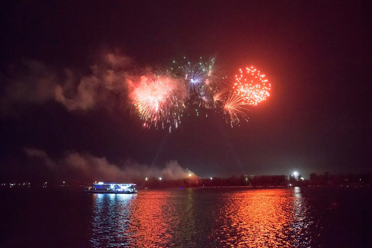 Fogos de artifício vermelhos e coloridos estourando no céu escuro sobre um rio, com o reflexo vermelho na água.