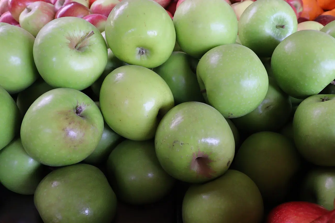 Maçãs verdes amontoadas numa banca de frutas, com algumas maçãs vermelhas e laranjas nos cantos.