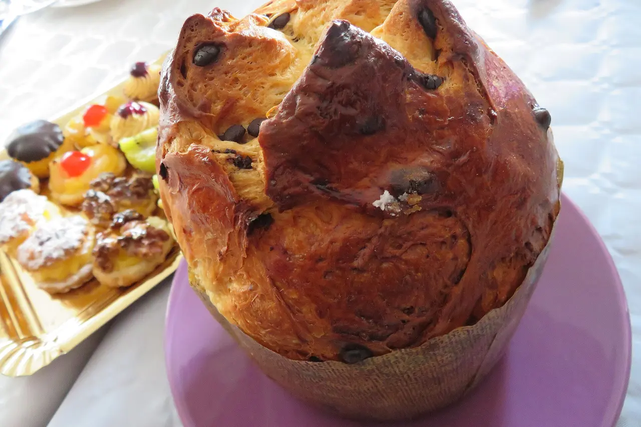 Panetone alto com gotas de chocolate num prato lilás, ao lado de uma bandeja dourada de docinhos.